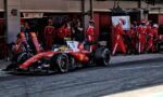 Red Ferrari Formula 1 car in the pit lane with crew members and a cameraman in the background.