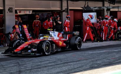 Red Ferrari Formula 1 car in the pit lane with crew members and a cameraman in the background.