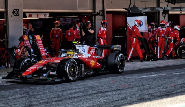 Red Ferrari Formula 1 car in the pit lane with crew members and a cameraman in the background.