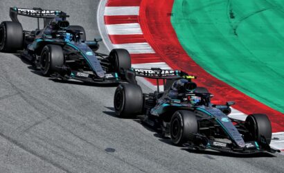 Three Mercedes AMG F1 cars in teal/black livery navigate a left-hand corner on a race track, red-and-white curb nearby and green run-off beyond.