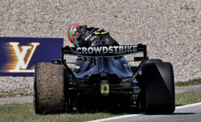 Open-wheel race car kicking up gravel on a track, front view with a colorful helmeted driver and CROWDSTRIKE on the rear wing, race banner in background.