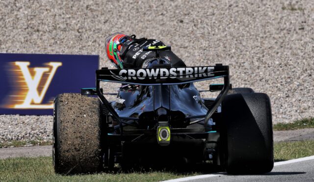 Open-wheel race car kicking up gravel on a track, front view with a colorful helmeted driver and CROWDSTRIKE on the rear wing, race banner in background.