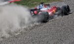 Red Formula 1 race car skids on a gravel runoff, kicking up a large cloud of dust and pebbles behind it on track limits.