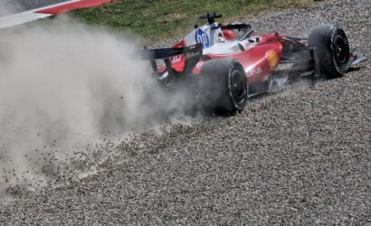 Red Formula 1 race car skids on a gravel runoff, kicking up a large cloud of dust and pebbles behind it on track limits.