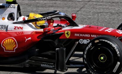 Ferrari Formula 1 car on track with driver in yellow helmet visible in the cockpit, front wing and red body covered in sponsor logos like Shell and ZYN