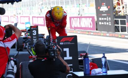 Racing driver in red suit and yellow helmet bows over a pit-stop podium with photographers and team equipment nearby.