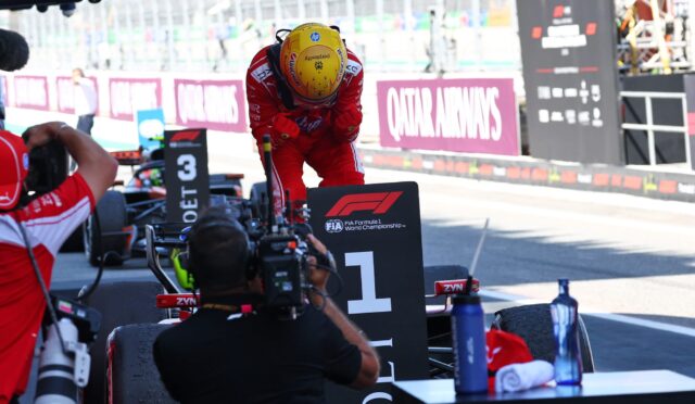 Racing driver in red suit and yellow helmet bows over a pit-stop podium with photographers and team equipment nearby.