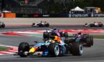 Formula 1 cars race through a chicane on a sunny track; the foreground car features blue and yellow Red Bull livery leading, with a pink/purple car close behind and others in the background by the barriers.