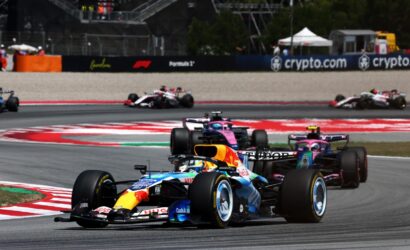 Formula 1 cars race through a chicane on a sunny track; the foreground car features blue and yellow Red Bull livery leading, with a pink/purple car close behind and others in the background by the barriers.