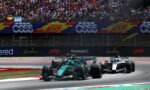 Two Formula 1 cars race a tight corner as a packed grandstand watches from behind colorful banners and fencing.