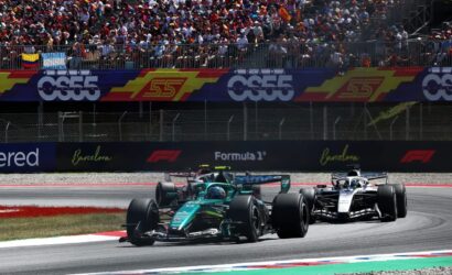 Two Formula 1 cars race a tight corner as a packed grandstand watches from behind colorful banners and fencing.
