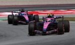 Two pink Formula 1 cars racing side by side on a track with a red and white barrier in the background.