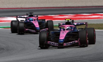 Two pink Formula 1 cars racing side by side on a track with a red and white barrier in the background.