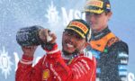 Racing drivers celebrate on the podium, spraying champagne as snow falls, wearing red suits and sponsor caps.