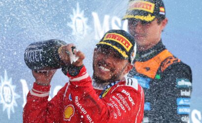 Racing drivers celebrate on the podium, spraying champagne as snow falls, wearing red suits and sponsor caps.