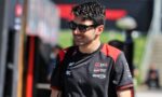 Man wearing black sunglasses and a black-red racing jersey, smiling outdoors with sponsor logos visible on the chest.