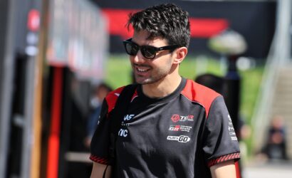 Man wearing black sunglasses and a black-red racing jersey, smiling outdoors with sponsor logos visible on the chest.