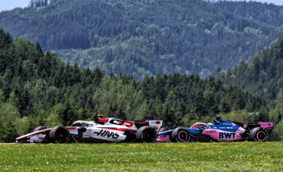 Row of colorful Formula 1 race cars lined up on a grassy field with a forested hillside in the background