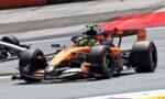 Orange Formula 1 car racing on track with a lime-green helmeted driver, close to a second car, red curb in the background.",