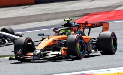 Orange Formula 1 car racing on track with a lime-green helmeted driver, close to a second car, red curb in the background.",