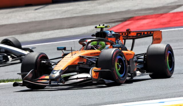 Orange Formula 1 car racing on track with a lime-green helmeted driver, close to a second car, red curb in the background.",