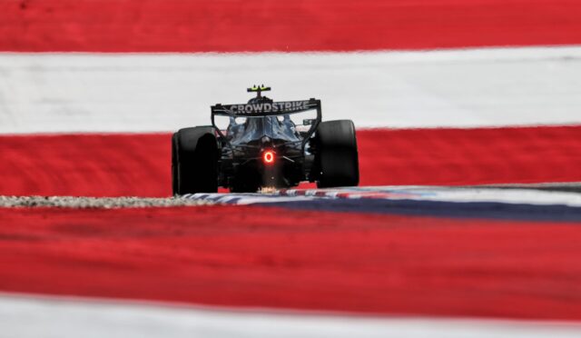 Rear view of a Formula 1 car racing on a track, red-and-white barriers in the background, CrowdStrike logo on the wing, red tail light visible