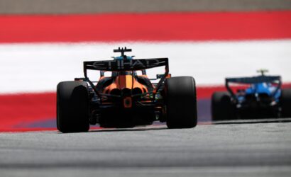 Rear view of an orange Formula 1 car on track, with Etihad branding on the rear wing and a second car blurred in the background.