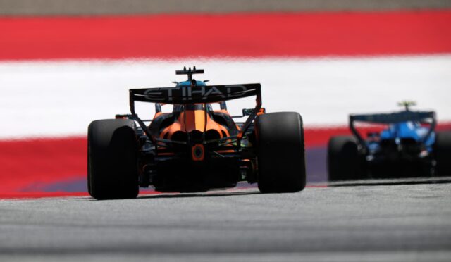 Rear view of an orange Formula 1 car on track, with Etihad branding on the rear wing and a second car blurred in the background.
