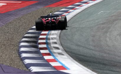 Red Formula 1 race car negotiating a sharp corner, hugging striped red, white, and blue curbs on the track.
