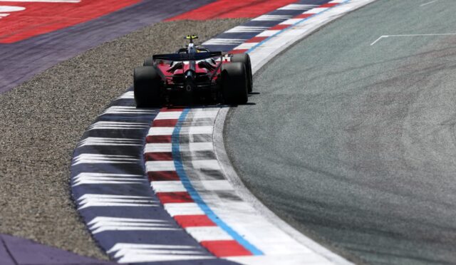 Red Formula 1 race car negotiating a sharp corner, hugging striped red, white, and blue curbs on the track.