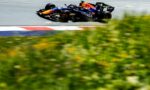 Red Bull Formula 1 car in mid-air over a race track, sponsor logos visible, motion blur on the foreground grass and barriers behind.