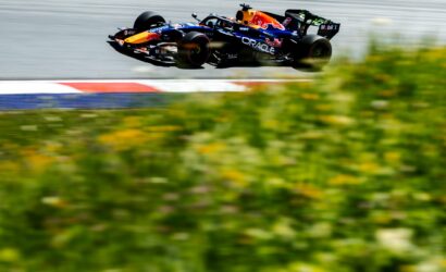 Red Bull Formula 1 car in mid-air over a race track, sponsor logos visible, motion blur on the foreground grass and barriers behind.