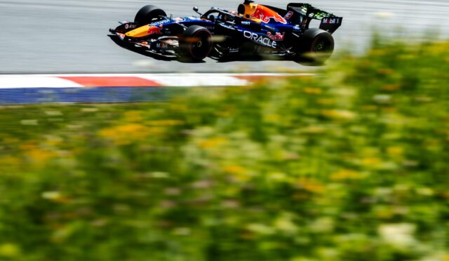 Red Bull Formula 1 car in mid-air over a race track, sponsor logos visible, motion blur on the foreground grass and barriers behind.