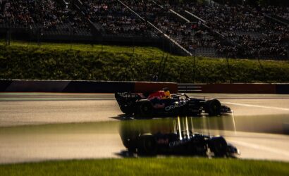 Formula 1 car with Red Bull livery racing along the track as grandstands full of spectators loom in the background.