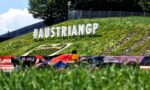 Large white 3D letters reading '#AUSTRIANGP' on a grassy slope beside a race track, with a Red Bull–style race car in the foreground.