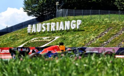 Large white 3D letters reading '#AUSTRIANGP' on a grassy slope beside a race track, with a Red Bull–style race car in the foreground.