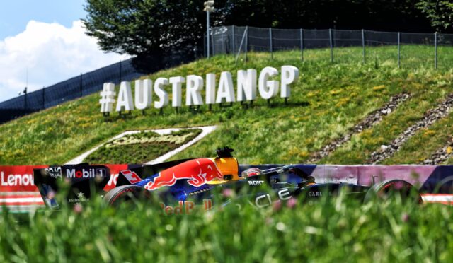Large white 3D letters reading '#AUSTRIANGP' on a grassy slope beside a race track, with a Red Bull–style race car in the foreground.