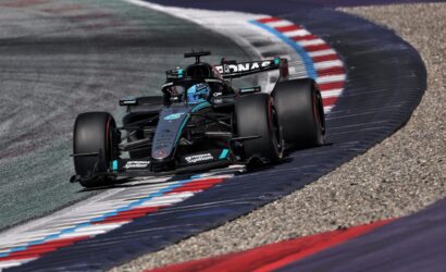 Mercedes F1 racing car navigating a sharp left turn on a track with red, white, and blue curbs.