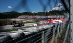 Formula 1 car on a curving race track with grandstands full of spectators and forest backdrop, viewed through a metal barrier and tires in the foreground.