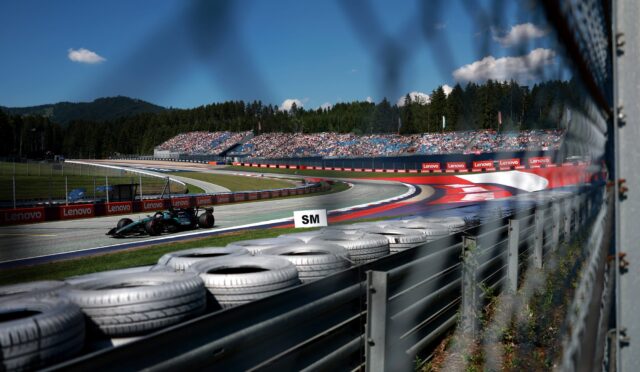 Race car on a winding track with tire barriers in the foreground, packed grandstands, and a forested hillside under a clear blue sky.