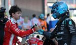 Two race drivers shake hands before a race, one in red suit and helmet, the other in a blue helmet and dark racing suit, in a pit area.