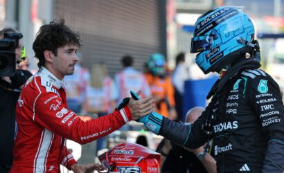 Two race drivers shake hands before a race, one in red suit and helmet, the other in a blue helmet and dark racing suit, in a pit area.