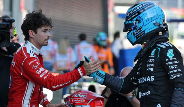 Two race drivers shake hands before a race, one in red suit and helmet, the other in a blue helmet and dark racing suit, in a pit area.