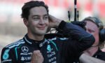 Smiling race car driver in a Mercedes-AMG racing suit celebrates with fists raised, background blurred.
