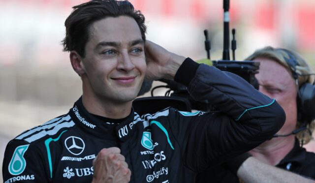 Smiling race car driver in a Mercedes-AMG racing suit celebrates with fists raised, background blurred.