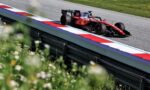 Red Formula 1 car on a race track beside red, white, and blue barriers, with green grass in the background and foreground flowers partially blurred.