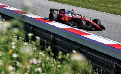 Red Formula 1 car on a race track beside red, white, and blue barriers, with green grass in the background and foreground flowers partially blurred.