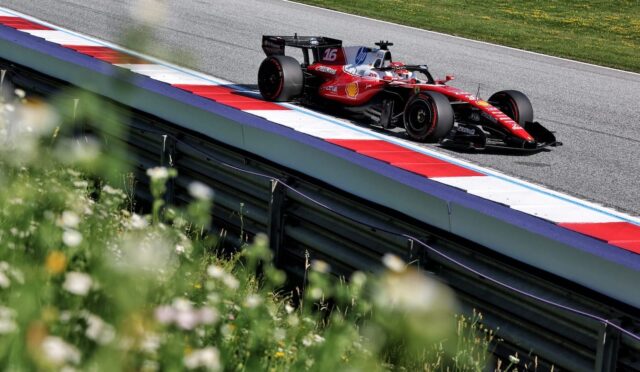 Red Formula 1 car on a race track beside red, white, and blue barriers, with green grass in the background and foreground flowers partially blurred.