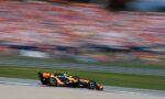 Orange Formula 1 car racing along a track with a crowded, blurred grandstand in the background (motion blur).