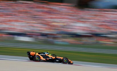 Orange Formula 1 car racing along a track with a crowded, blurred grandstand in the background (motion blur).